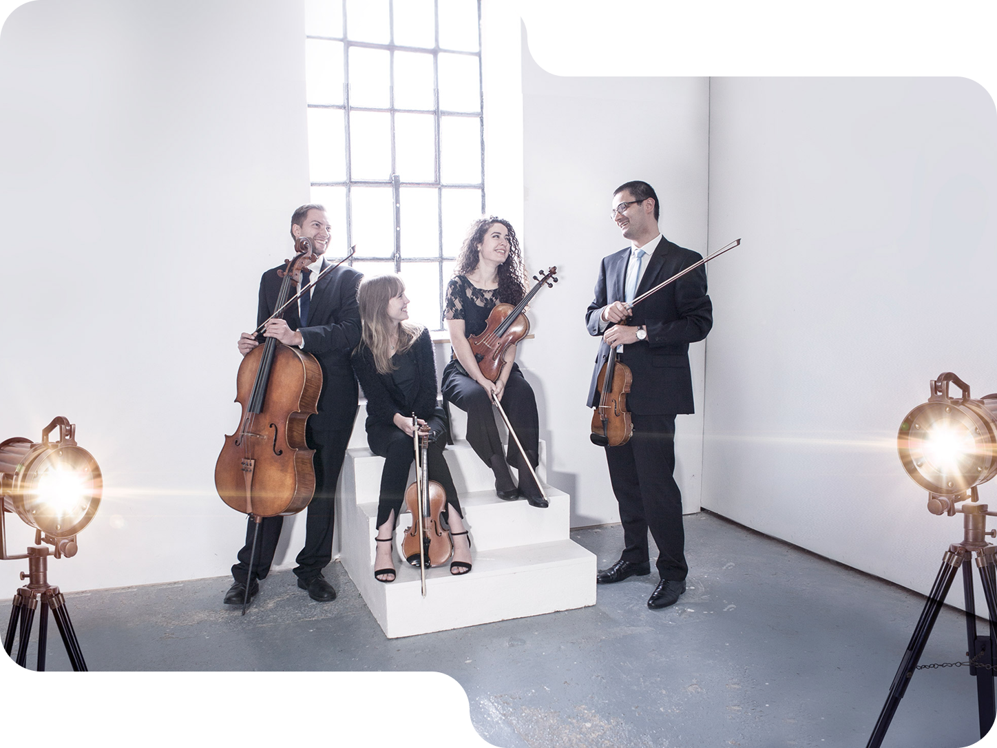 Four musicians dressed formally with string instruments posing in a bright white room with two studio lights.